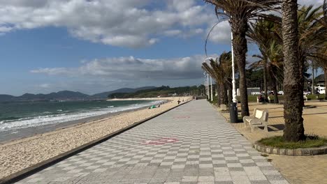 Pov-walk-along-promenade-of-Vigo-CIty-with-palm-trees,-sandy-beach-and-strong-ocean-waves