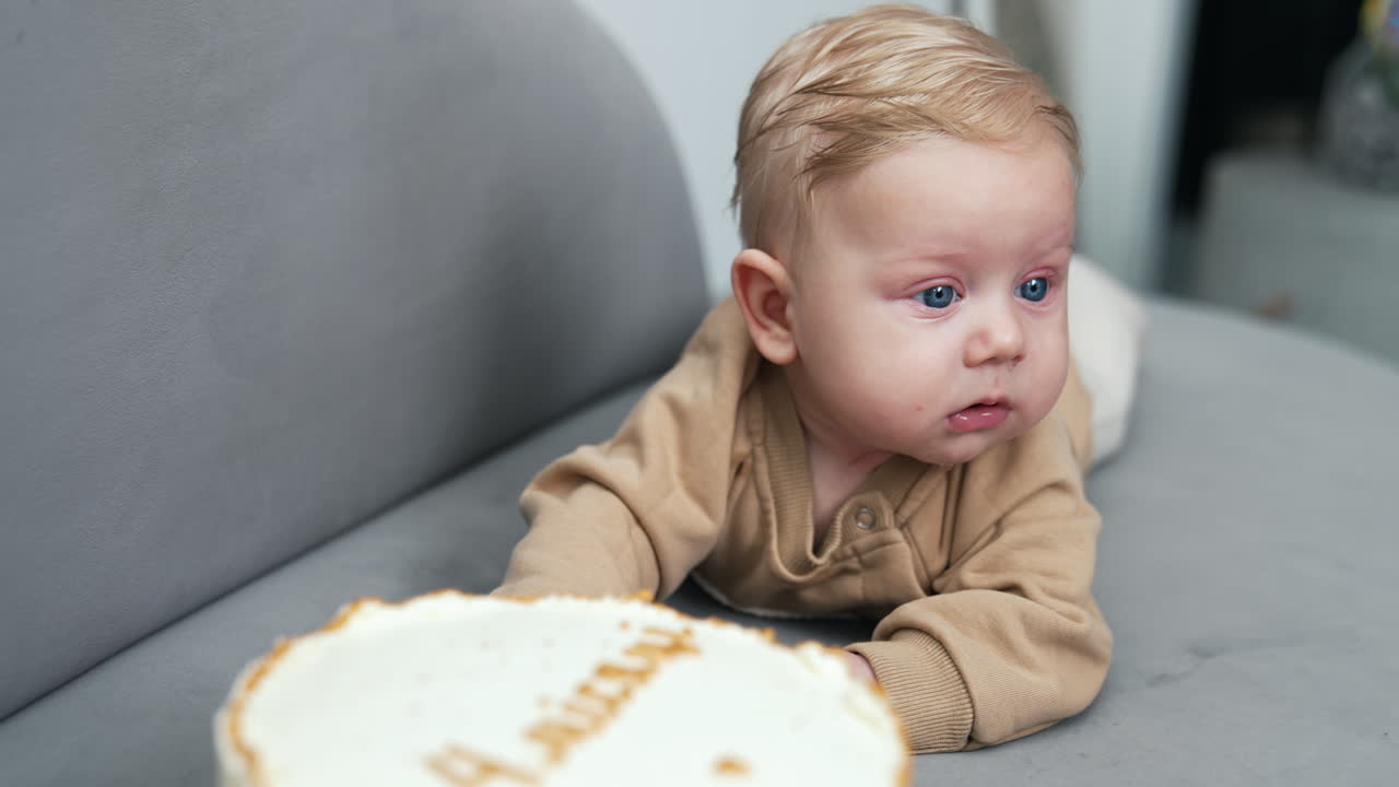 Beautiful adorable Caucasian baby lying near a big cake. Calm blond infant lies on belly looking around.