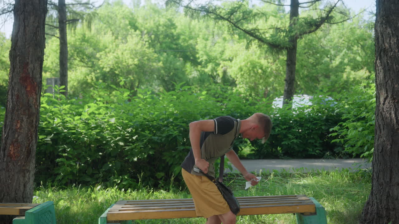 Young White Man Searching Bag On Bench Under Trees, Tense Posture And Quick Glances, Backpack Strap In Hand, Summer Sunlight Through Foliage, Solitary Urban Park Scene With Nervous Fidgeting