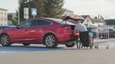 adult man with disabilities in a wheelchair puts purchases in the trunk of a car in a supermarket parking lot