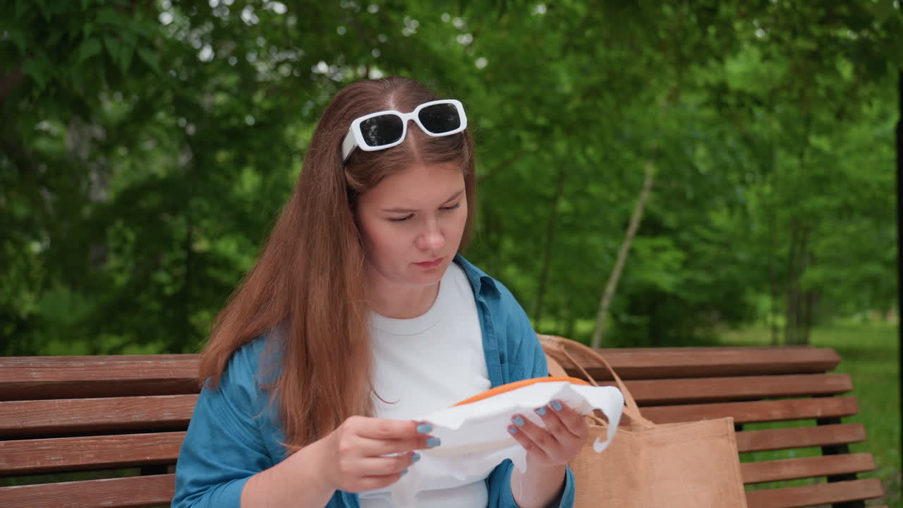 Una joven sentada en un banco del parque observa de cerca un bordado mientras señala el diseño con tristeza, rodeada de árboles verdes y una suave luz solar.