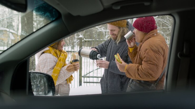 Tre amici che bevono e mangiano un panino in piedi vicino all'auto durante un viaggio in auto invernale