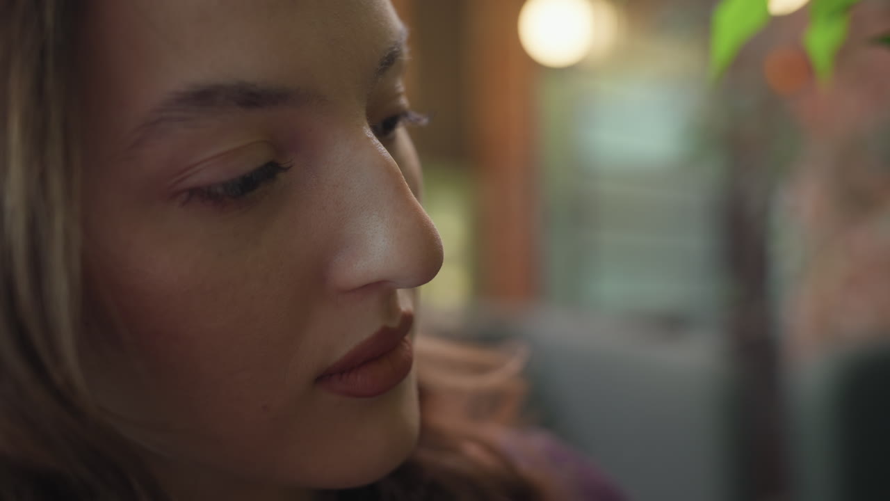 Close up face of woman with brown eyes and pointed nose looking upward in soft ambient light with blurred background, showing serene expression and warm tones across face and lips in indoor setting