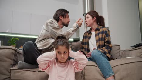 una pequeña chica morena con ropa rosa se cubre las orejas y está triste mientras sus padres están peleando mientras están sentados en el sofá en un apartamento moderno. un hombre con paja en una camiseta beige está peleando con su esposa morena en una camisa a cuadros mientras su hija pequeña está sentada en el suelo y cubriendo