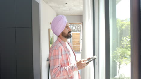 using tablet, man in turban standing by window in modern home