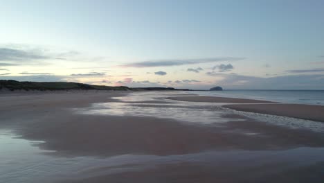 slow moving drone footage flying close to the surface of a long sandy beach before rising up high above it towards a sunset while the tide laps the shore