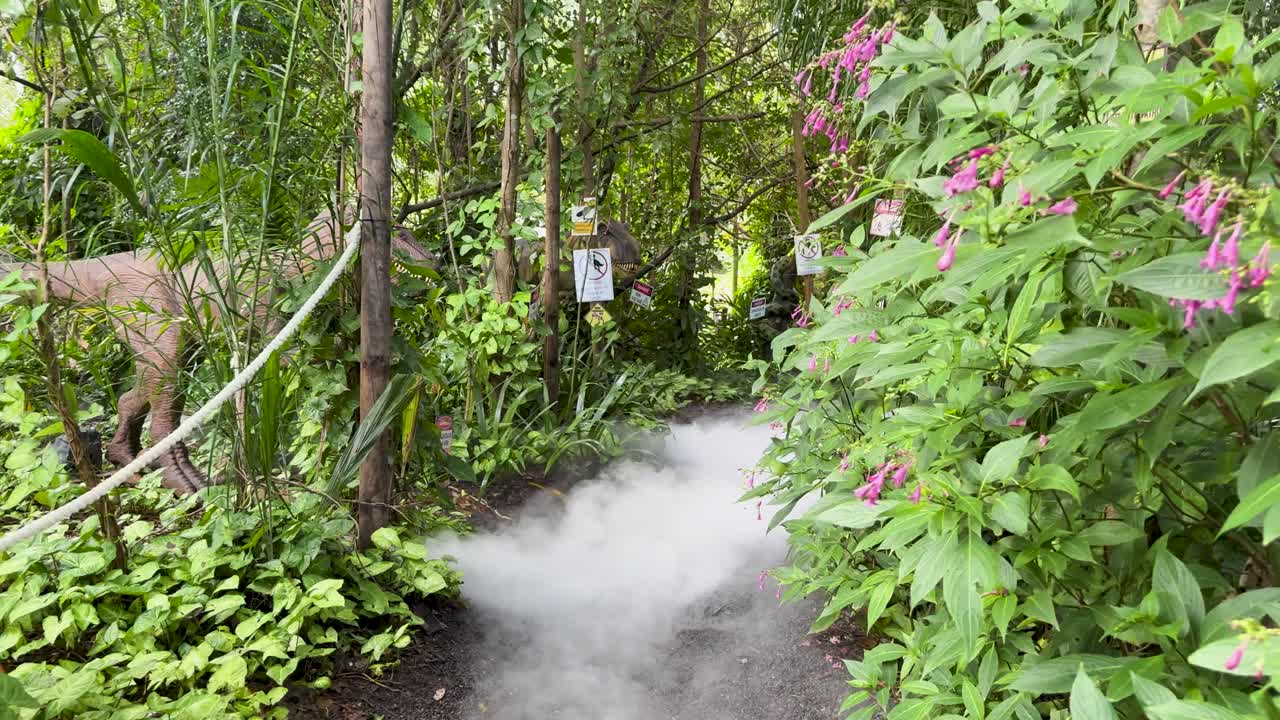 Animatronic dinosaur tail moves beside misty jungle path, lush greenery, natural daylight, static camera