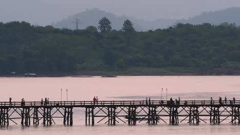 Mon-Bridge-and-a-longboat-followed-speeding-behind-the-structure-while-people-are-walking-on-the-bridge,-silhouetting-as-it-was-getting-dark,-in-slow-motion