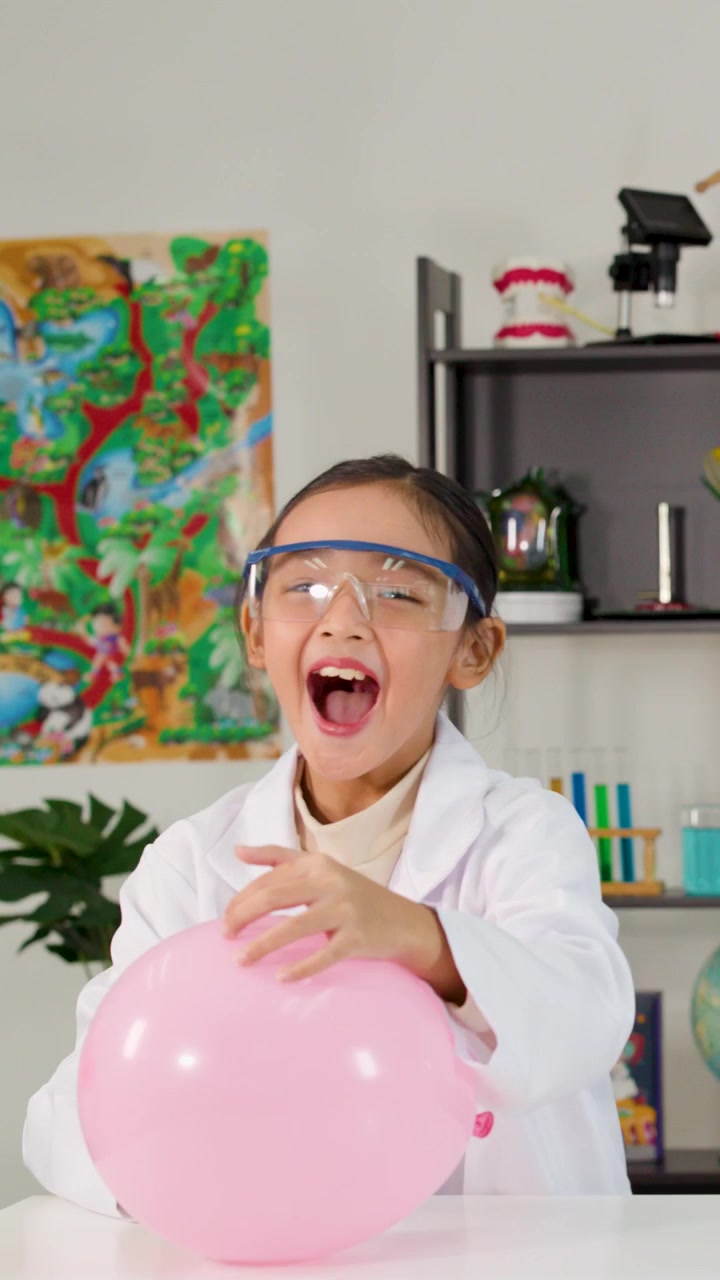 Young students in lab coats and goggles laugh while popping balloons during a classroom experiment