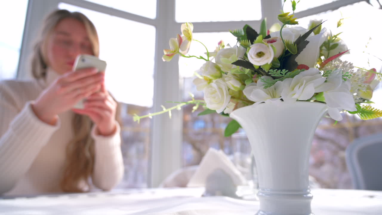 レストランのテーブルで白い花を前に、スマホをスクロールする女性。