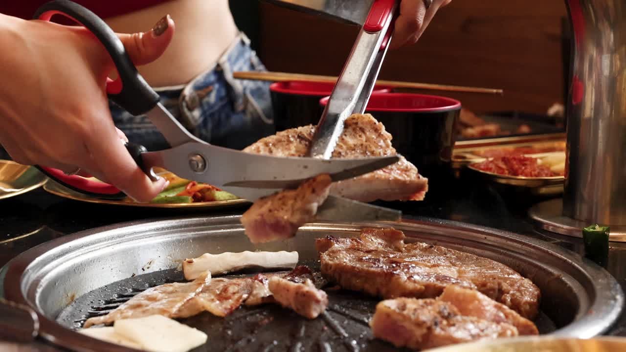 Person uses scissors to cut pork belly on tabletop Korean barbecue grill, warm ambient lighting