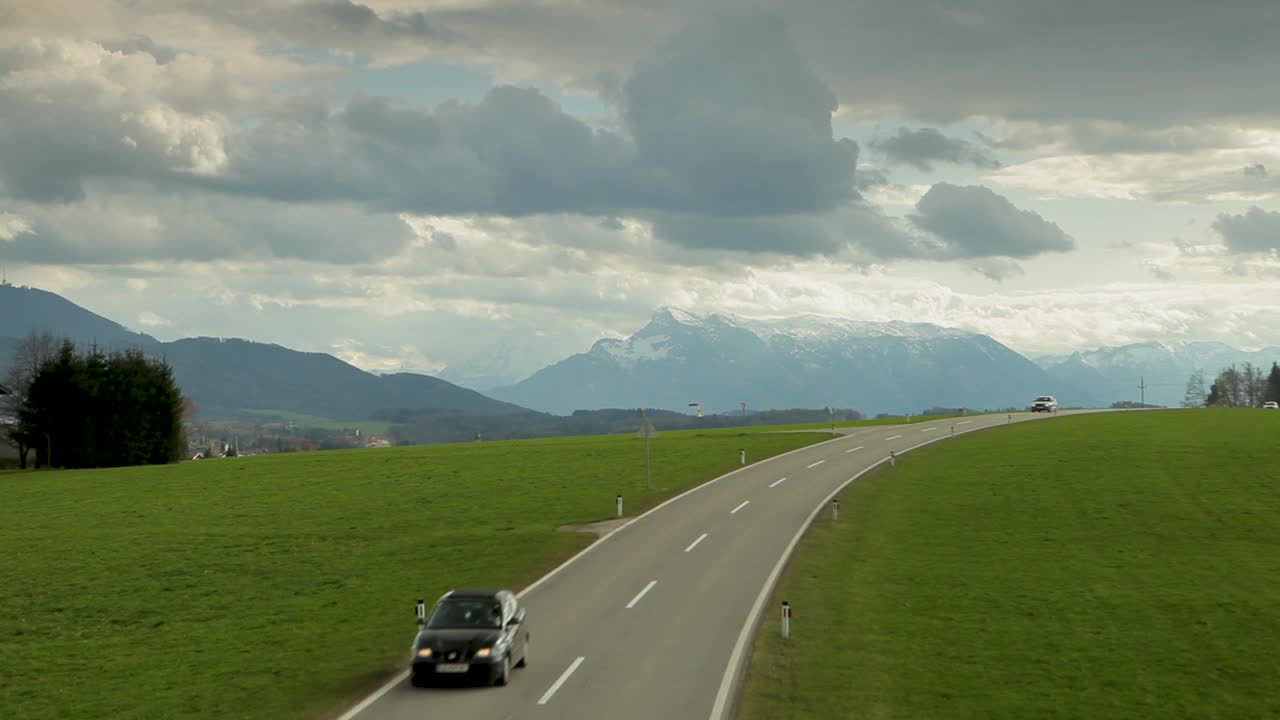 자동차는 초록색 과 눈 인 산을 배경으로 고속도로를 운전합니다. 하늘은 부분적으로 구름이 많습니다.
