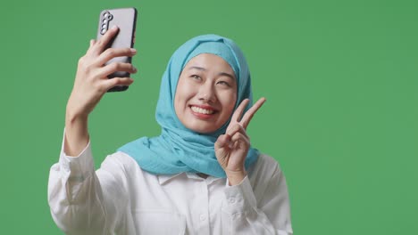 close up of asian muslim woman smiling and using smartphone taking picture while standing in the green screen background studio