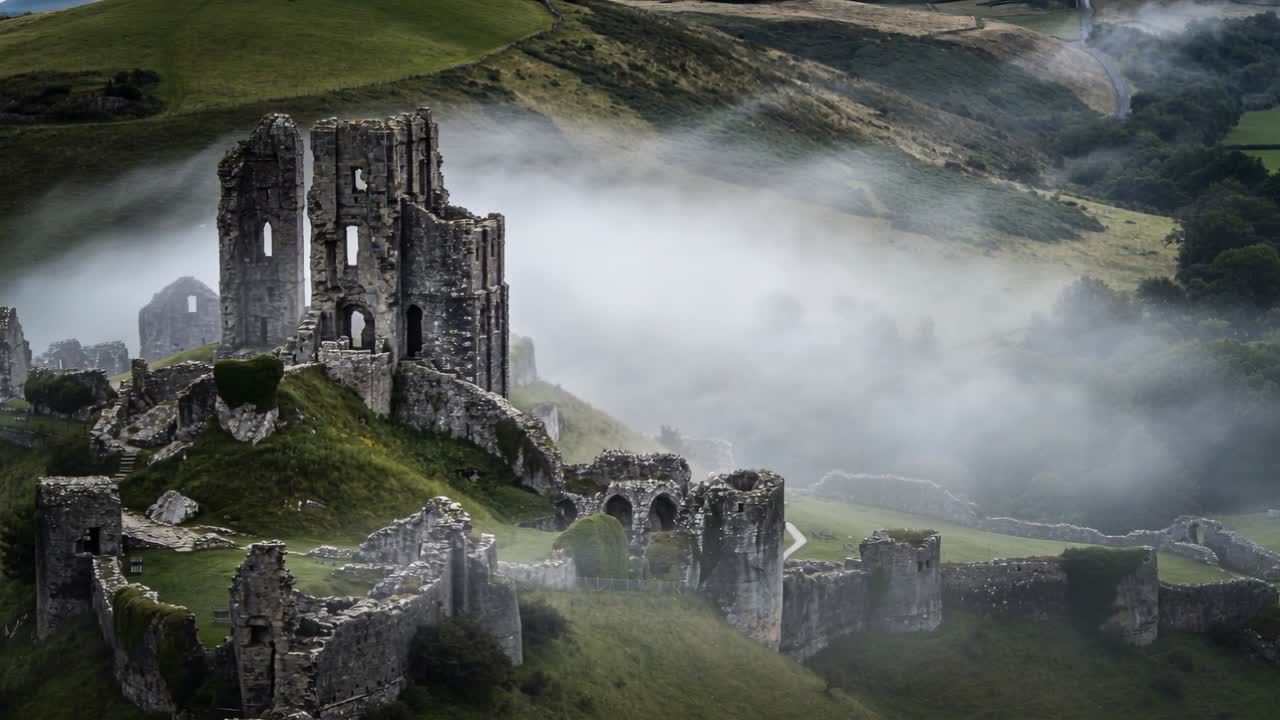 La telecamera zooma, enfatizzando un castello di pietra in rovina in cima a una collina erbosa, con la nebbia che si sposta verso destra.