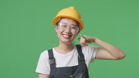 close up of asian woman worker wearing goggles and safety helmet smiling and making call me gesture to camera while standing in the green screen background studio