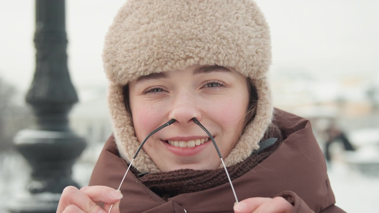 Close frontal view of young woman outdoors in cold winter weather, lifting then lowering glasses while speaking with slight laugh and visible breath, showing casual urban lifestyle mood