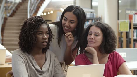 Señoras-Sonrientes-Hablando-Mientras-Están-Sentadas-En-La-Mesa-Con-Una-Laptop-En-La-Biblioteca