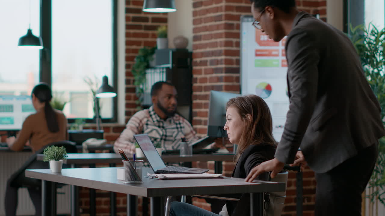 Young man helping businesswoman in wheelchair to work at desk