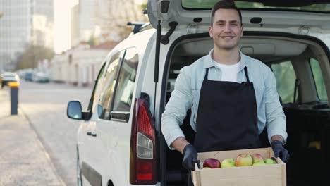 happy smiling man with braces holding wooden box with apples at street outdoors, portrait of happy delivery man in apron and