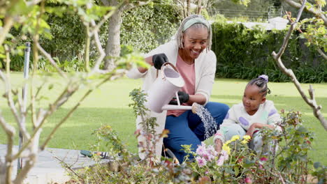 Feliz-Abuela-Y-Nieta-Afroamericana-Regando-Plantas-En-Un-Jardín-Soleado,-Cámara-Lenta