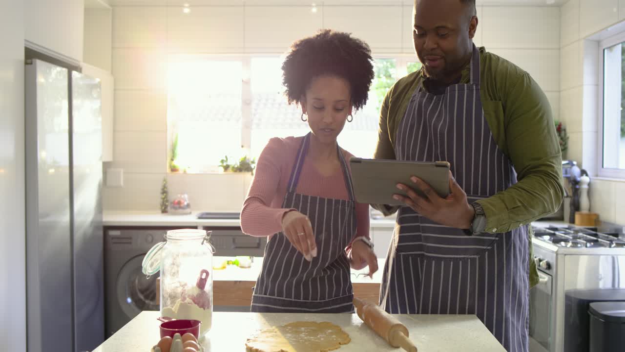 Diverse couple entering kitchen using tablet reading recipe while woman flouring dough, rolling