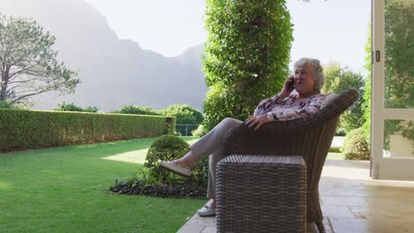 caucasian senior woman smiling while talking on smartphone while sitting on the chair outdoors