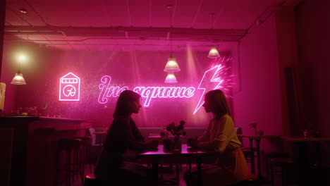 two women engaged in conversation at a neon-lit cafe