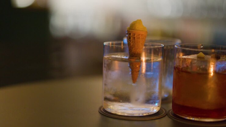 Bartender places small cone garnish in cocktail glass with ice, warm bokeh lighting, shallow focus