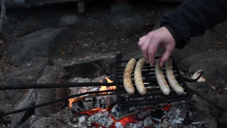 White-hand-changing-grilling-sausage-bratwursts-side-on-grill-net-over-open-camp-fire