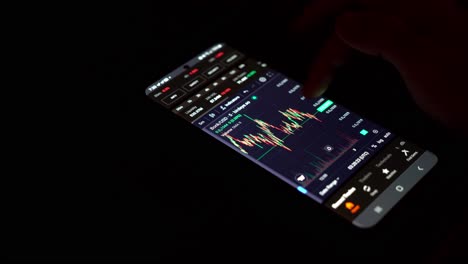 Person-scrolling-through-cryptocurrency-charts-on-a-smartphone-in-a-dark-setting