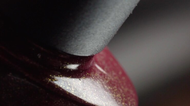 Primo piano macro di un flacone di smalto per unghie con tappo nero e lacca rossa lucida intorno al collo del flacone, che rivela i dettagli della confezione cosmetica con messa a fuoco selettiva e illuminazione soffusa