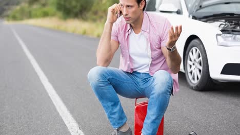 hombre caucásico sentado en un bote hablando en un teléfono inteligente por un coche averiado en una carretera rural