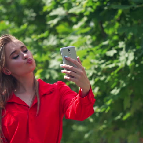 pretty girl doing selfie outdoors. sexy woman posing on her cell phone on the green tree background. beautiful female with long hair in red dress holds mobile phone.