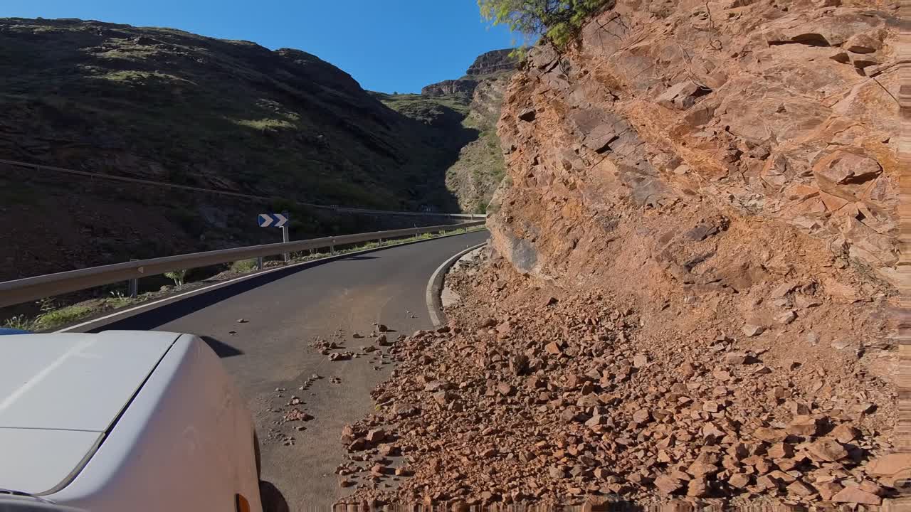 Riprese POV di un veicolo che percorre una pericolosa strada di montagna parzialmente bloccata da una frana. Rocce e detriti coprono la strada asfaltata.