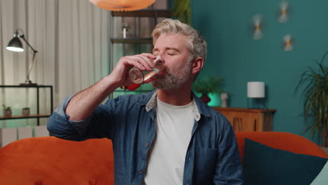 thirsty middle-aged man sitting at home holding glass of natural aqua make sips drinking still water