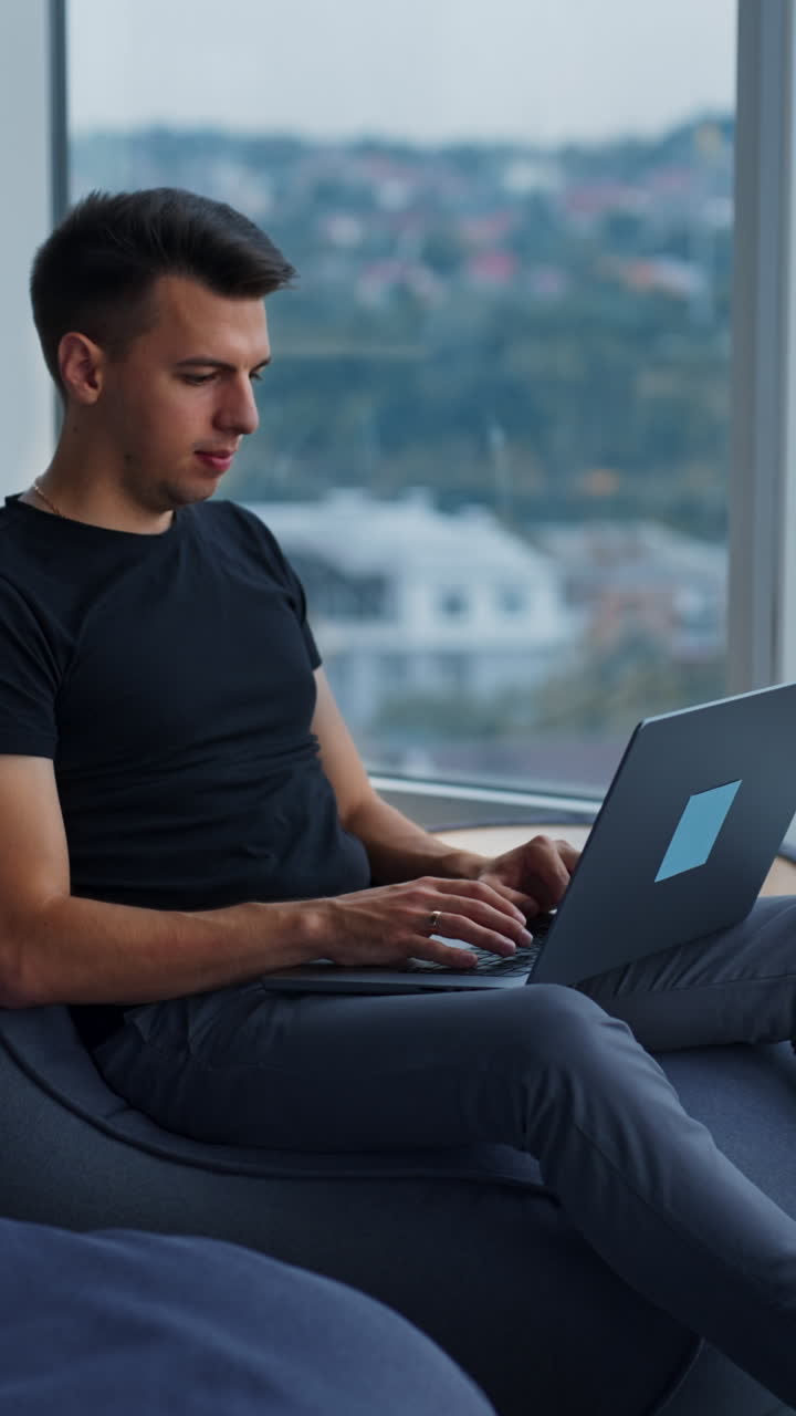 Young male freelancer working on laptop sitting comfortably near the panoramic window. Side view. Cityscape at backdrop in blur. Vertical video