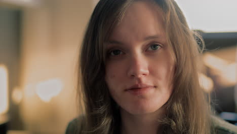 Close-up-shot-of-young-white-Woman-looking-at-herself-into-the-camera-and-smiling-during-golden-hour-sunrise-in-her-apartment-in-slow-motion