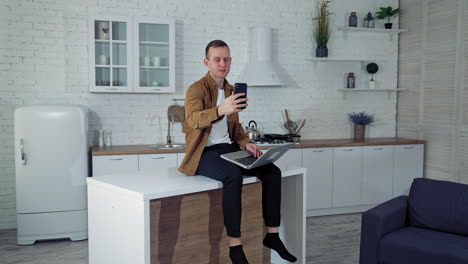 young man working online at home. freelance male sitting on a kitchen table with a laptop and having a video call through the phone. distant job.