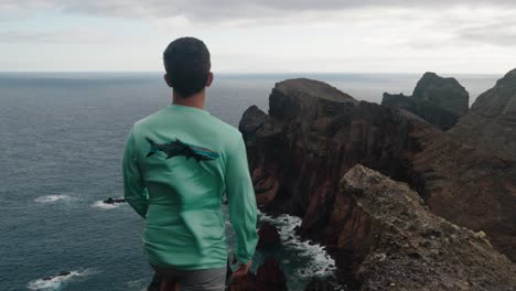 a man stands on a cliff edge overlooking dramatic red and black rock formations on the coast of madeira, portugal.
