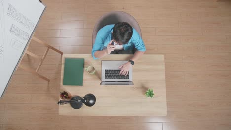 vista superior de un hombre hablando en un teléfono inteligente mientras está sentado en el escritorio de madera además de una pizarra escribiendo en una computadora portátil en casa