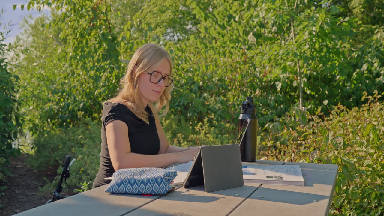 SchülerIn der Generation Z mit Brille, die draußen mit Tablet, Notizbuch und wiederverwendbarer Wasserflasche an einem Tisch im Park lernt. Dolly-Aufnahme, die sich nach rechts bewegt, Konzept von Lernen im Freien, Fokus, Wissen und Nachhaltigkeit