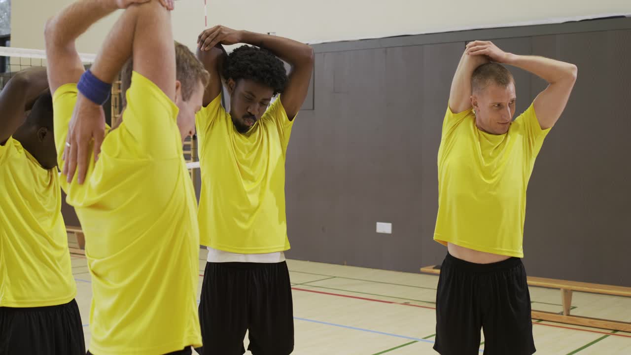 Six male teammates in yellow shirts performing shoulder triceps stretches in gym near net warming