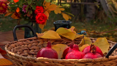 Granadas-Rojas-Maduras-En-Una-Cesta-Sobre-Una-Mesa-De-Jardín-Con-Un-Ramo-De-Flores-En-Un-Jarrón-Detrás