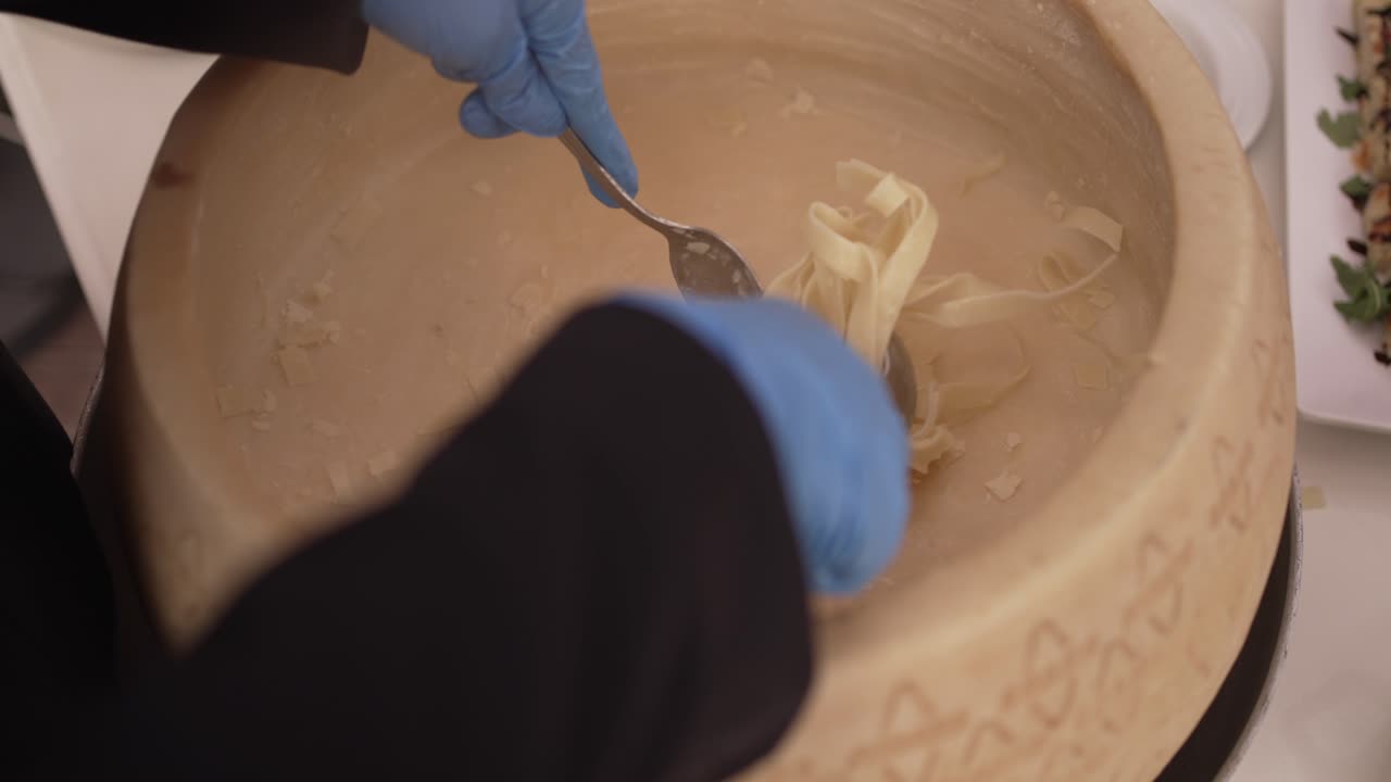 Pasta being mixed inside a large cheese wheel by a chef using utensils with gloved hands