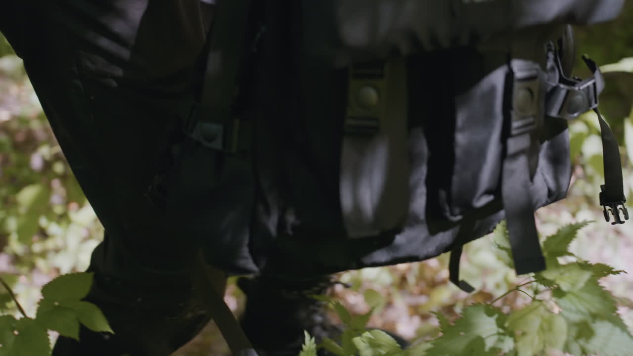 Side view of soldier walking through grassy forest terrain, pausing mid-step while turning leg to shake off debris, with sunlight casting shadows across black trousers and leafy ground cover