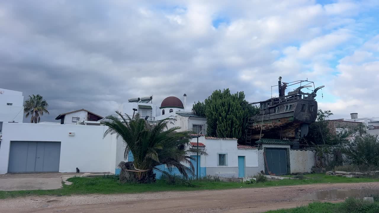 Inquadratura statica di una casa bianca con una palma e una grande struttura di nave in legno accanto, a Barbate, Cadice, sotto un cielo nuvoloso