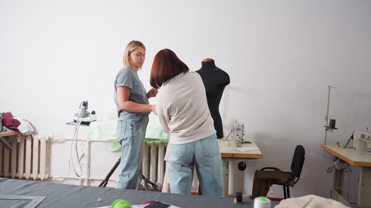 Two female tailors working together in workshop, one removing material from mannequin while other prepares it for ironing, electric pressing iron nearby, teamwork, fashion design activity