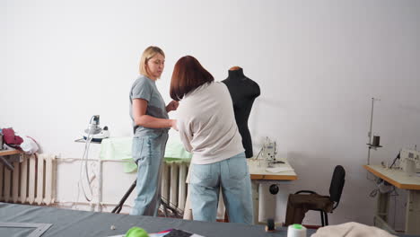 two female tailors working together in workshop, one removing material from mannequin while other prepares it for ironing, electric pressing iron nearby, teamwork, fashion design activity