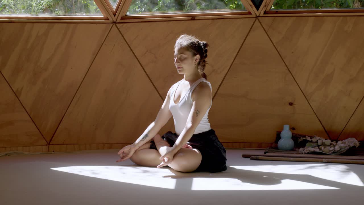 mujer en una pose de yoga pacífica dentro de un estudio al aire libre zen con formas geométricas, abrazando la atención plena, sentada con las piernas cruzadas
