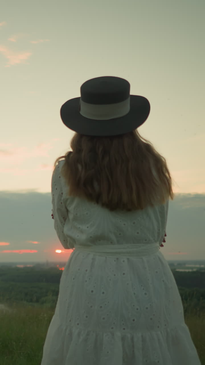 Close-up back view of a woman in a white dress and hat, arms folded, standing alone in a serene grassy field, gazing out over a tranquil lake during sunset, under a soft, pastel-colored sky